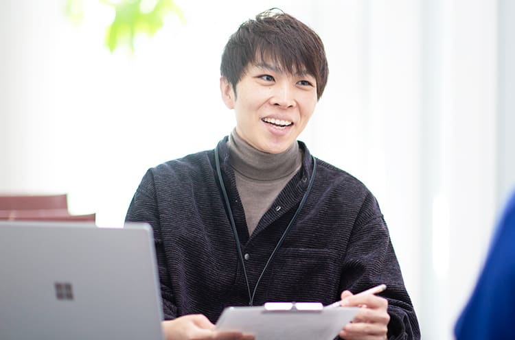 天間悠太さんが笑顔で相手の方と話をしながらクリップボードに書き込みをしている写真